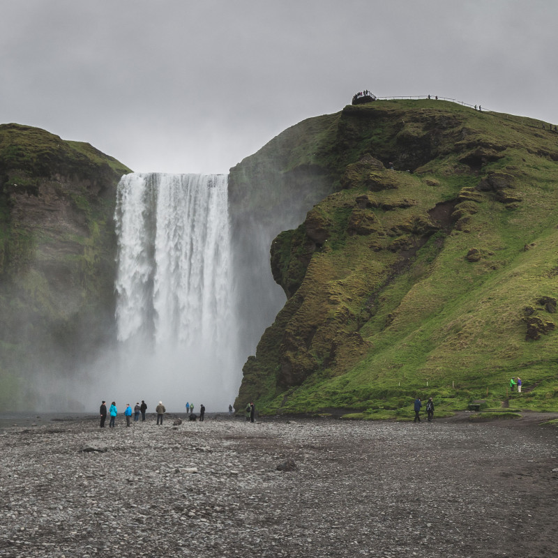 Wasserfall Skógafoss umringt von Bergen, im Vordergrund winzige Menschen.