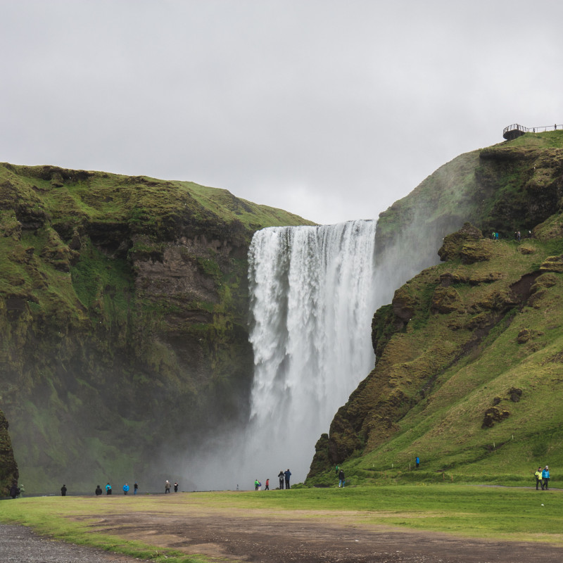 Wasserfall Skógafoss umringt von Bergen, im Vordergrund winzige Menschen.