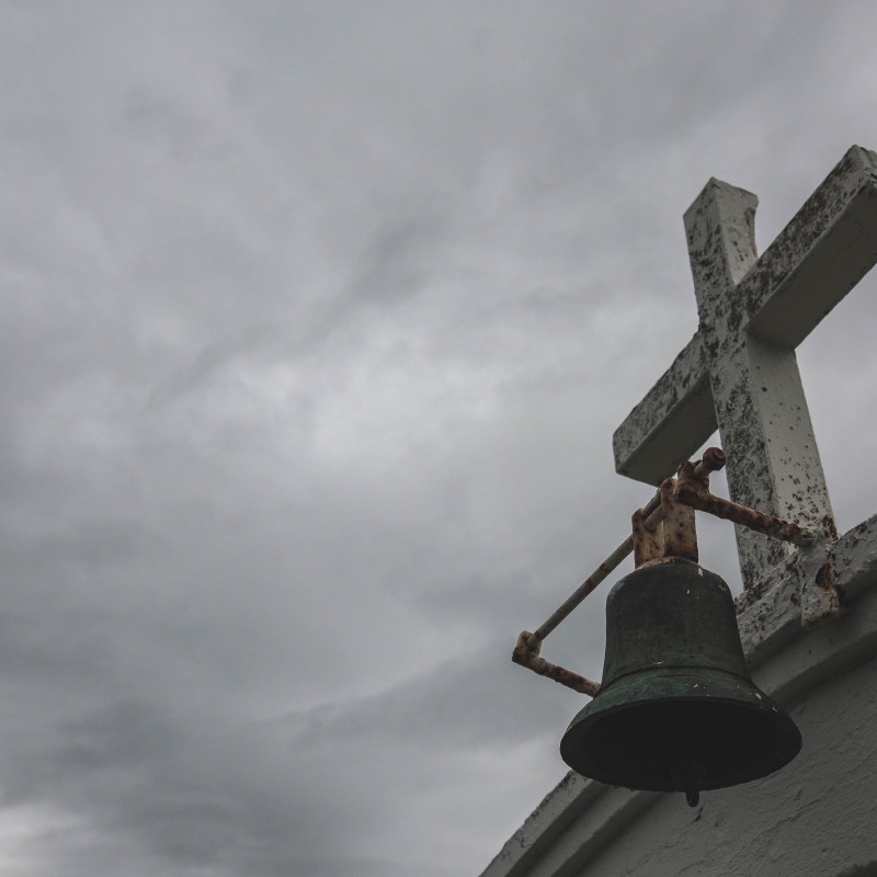 Kreuz und Glocke vor dichten grauen Wolken.