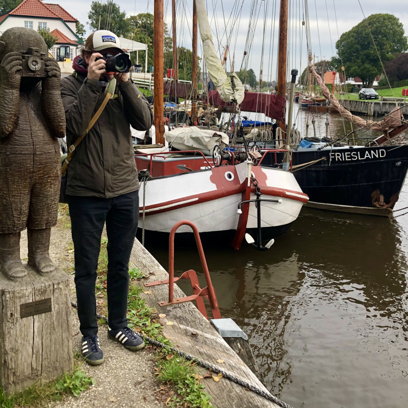 Ein Mann steht mit der Kamera neben einer Holzfigur die ebenfalls eine Kamera hält, im Hintergrund Schiffe.