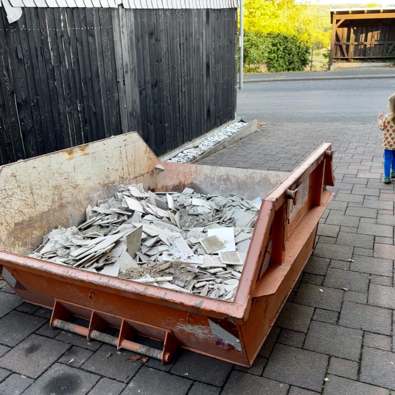 Ein orangener Container voller Bauschutt und ein Kind was uninteressiert vorbeiläuft. 