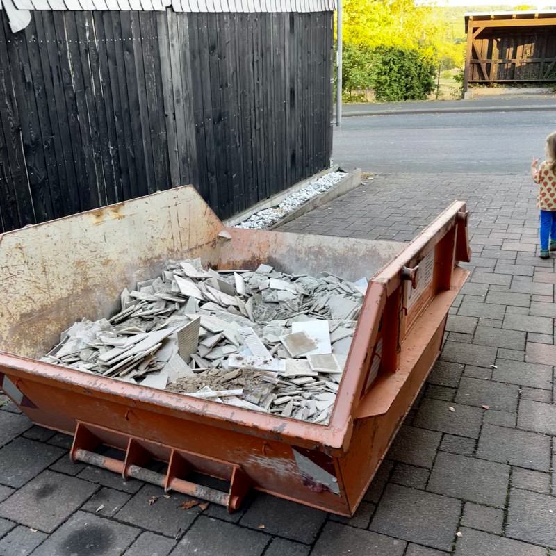 Ein orangener Container voller Bauschutt und ein Kind was uninteressiert vorbeiläuft.