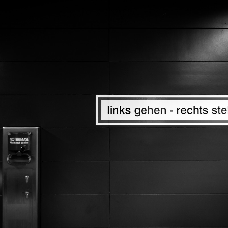 Schwarz-weiß-Foto: Ein Schild an einer Rolltreppe mit der Aufschrift "Links gehen, rechts stehen" und ein Hebel der mit "Notbremse" beschriftet ist.