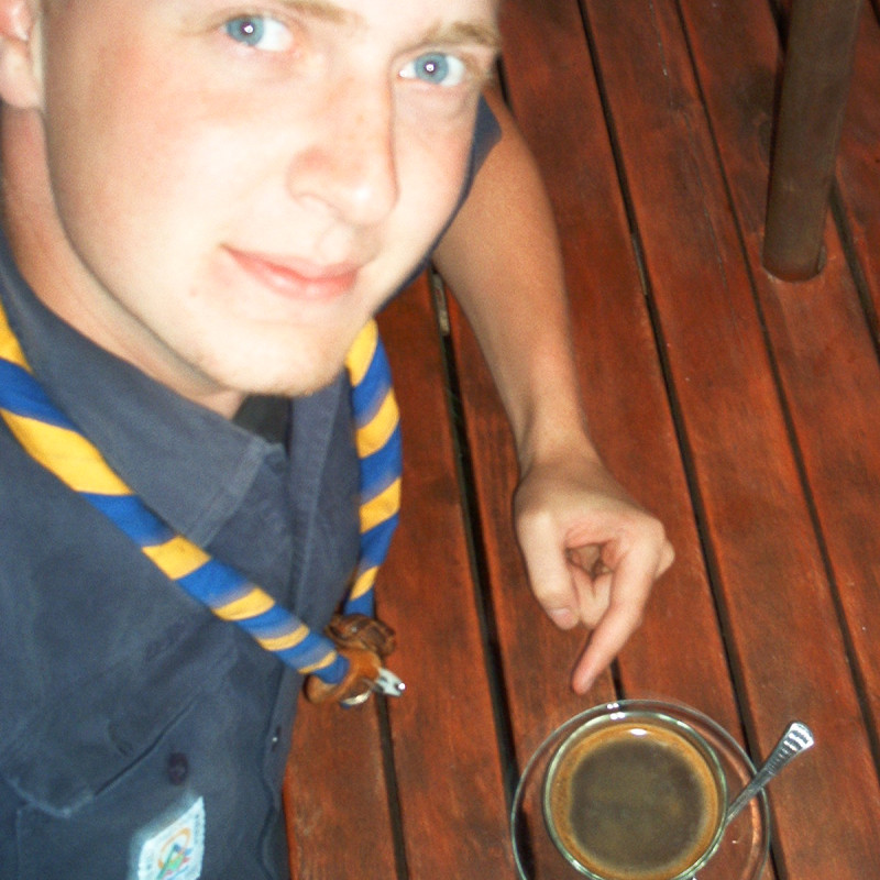 Martin schaut nach oben in die Kamera und zeigt auf einen Kaffee der vor ihm auf dem Tisch steht.