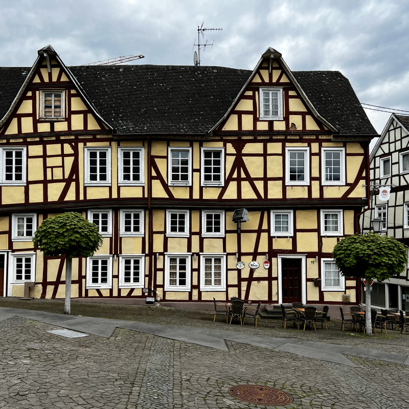 Ein breites gelbes Fachwerkhaus steht ein bisschen krum an einem Platz. Ringsherum weitere Fachwerkhäuser.