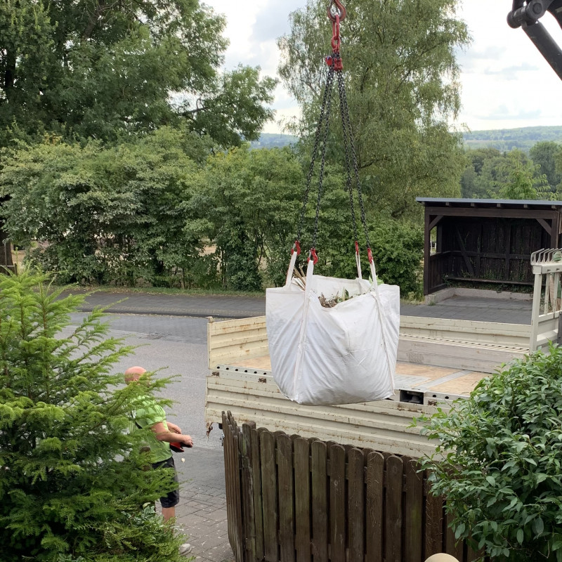 Ein großer weißer Sack voller Grüngut hängt an einem Kran und wird auf einen LKW verladen.