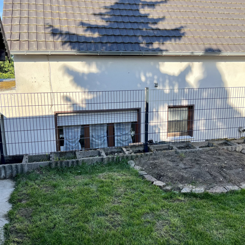 Ein Zaun steht vor einem Haus, im Vordergrund ein unbepflanztes Beet und eine Wiese.