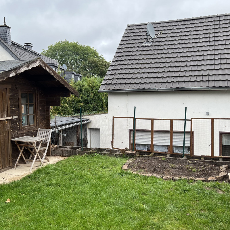 Eine Wiese mit einem leeren Beet, links ein braunes Gartenhaus. Im Hintergrund ein kaputter Zaun.