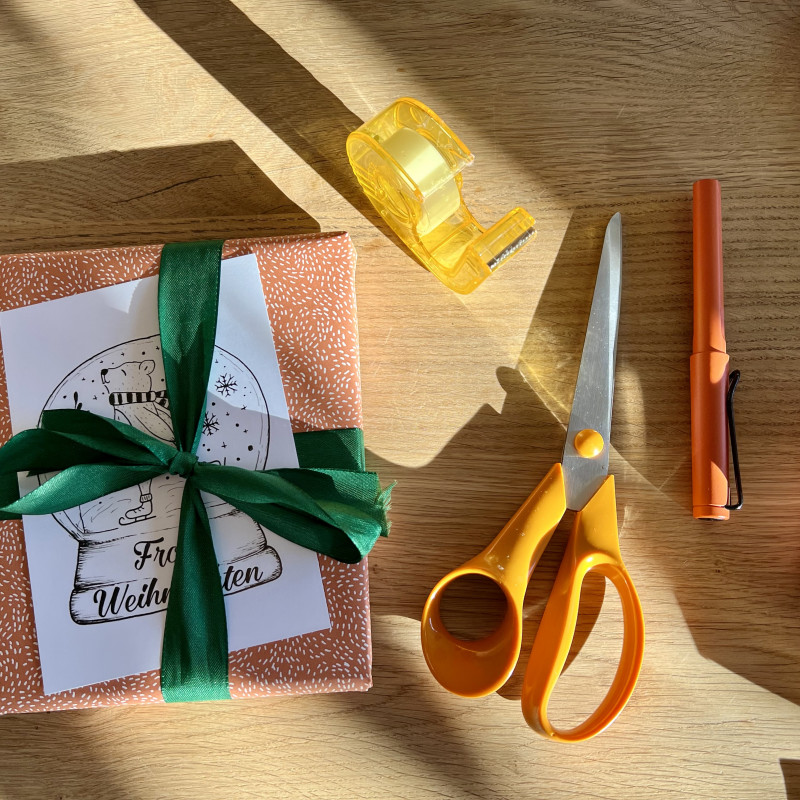 Ein in rot-weiß gepunktetes Geschenkpapier verpacktes Geschenk mit einer grünen Schleife, daneben Klebeband, eine Schere und ein Füller.
