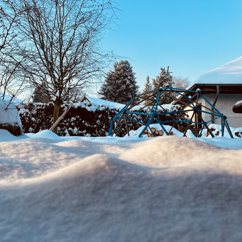 Blick in einen schneebedeckten Garten, vorne eine große Schneewehe, im Hintergrund ein Klettergerüst.