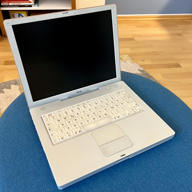 Ein weißer Laptop mit der Aufschrift iBook steht auf einem blauen Sessel.