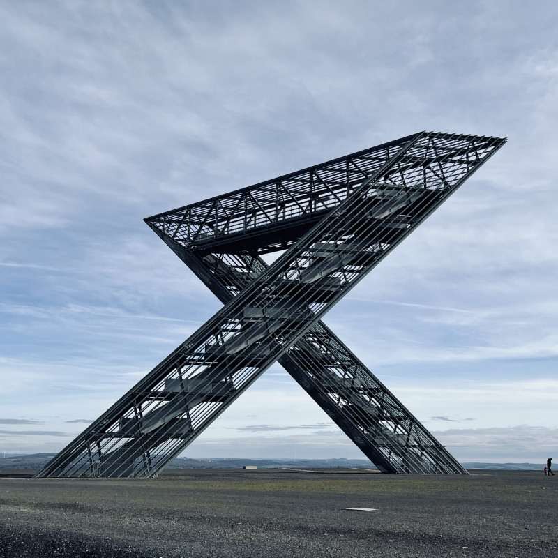 Das Saar-Polygon, eine begehbare Stahl-Skulptur vor blauem Himmel mit leichter Bewölkung. Ganz klein sind ein Mann und ein Kind zu sehen.