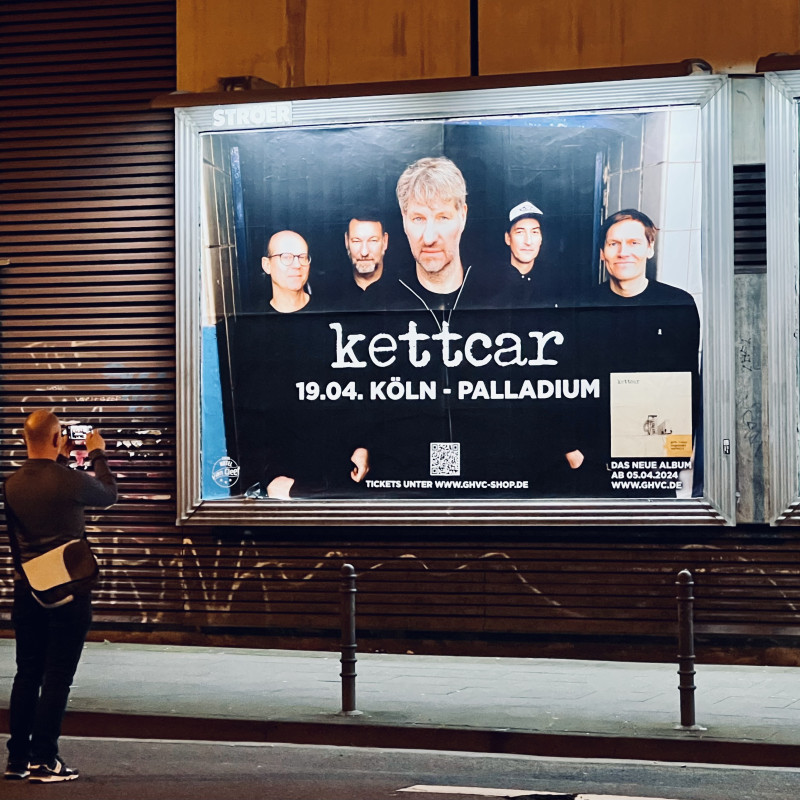 Ein großes Plakat mit Werbung für das Kettcar-Konzert am 18.4. im Palladium. Davor ein Mann der ein Foto mit seinem Mobiltelefon davon macht.
