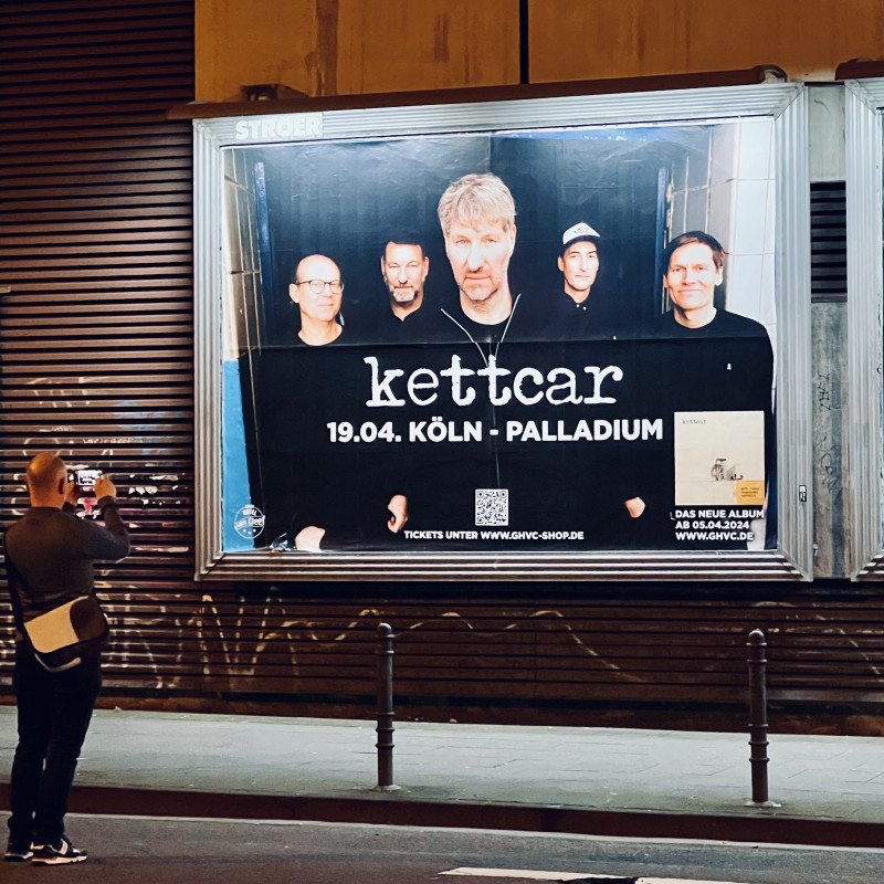 Ein großes Plakat mit Werbung für das Kettcar-Konzert am 18.4. im Palladium. Davor ein Mann der ein Foto mit seinem Mobiltelefon davon macht.