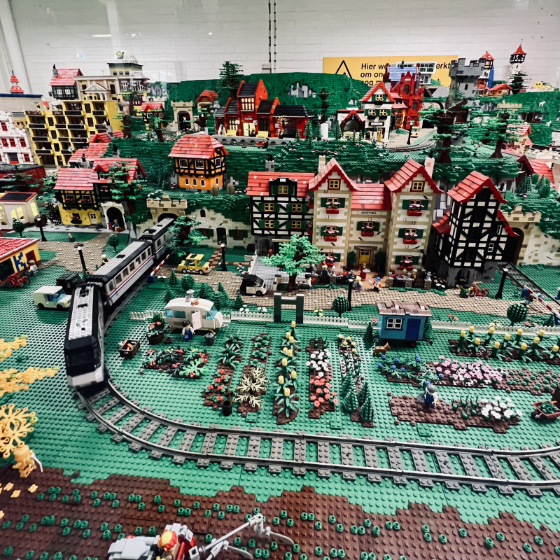 Blick auf eine aus LEGO-Steinen gebaute Stadt mit vielen Häusern, Blumen und Figuren. Im Vordergrund fährt ein Zug vorbei.
