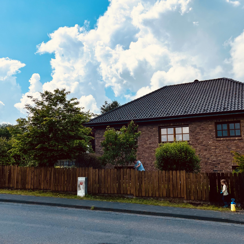 Ein Haus vor blauem Himmel mit Wolken, im Vordergrund ein Holzzaun.