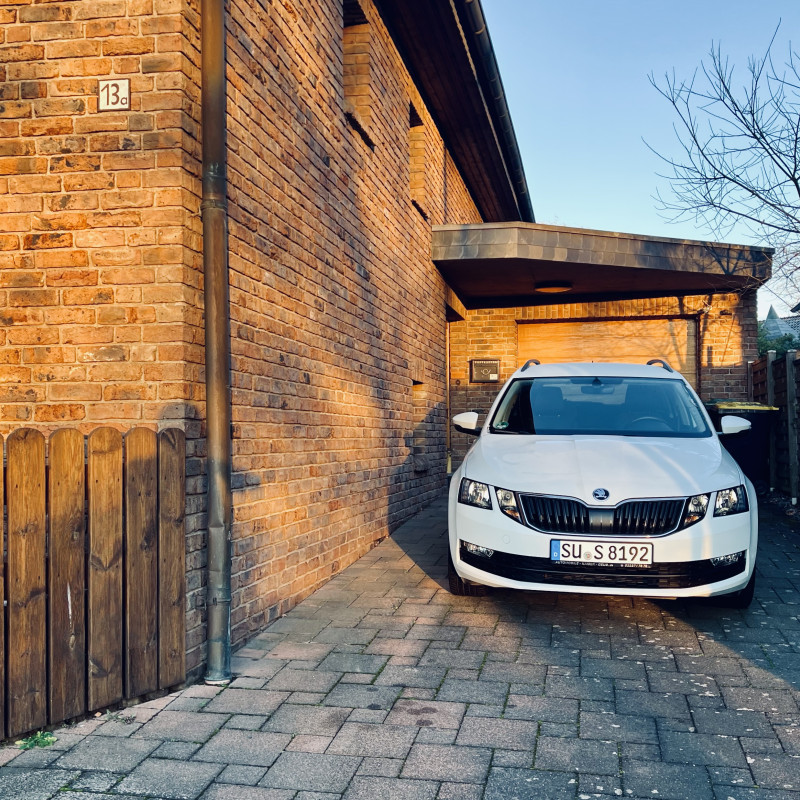 Ein weißes Auto steht im Morgensonnenschein in der Einfahrt neben einem verklinkerten Haus.