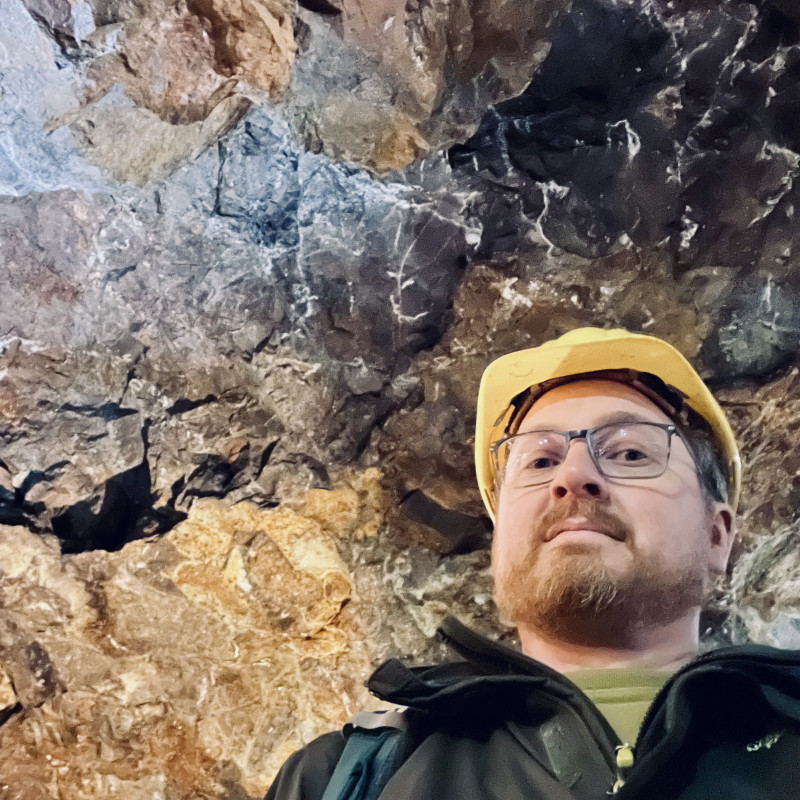 Ich blicke von oben in die Kamera, trage dabei einen gelben Schutzhelm. Im Hintergrund die grob behauenen Wände eines Bergwerks.