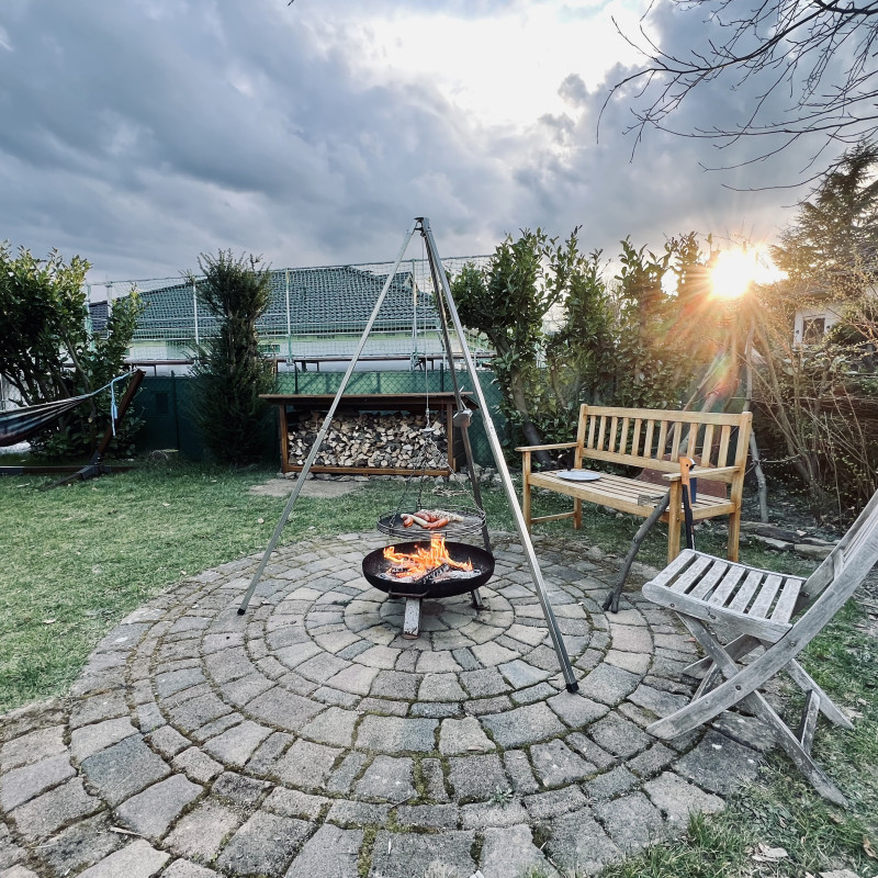 Über einem Feuer liegen rote und weiße Würstchen auf einem Grillrost, der an einem Dreibein hängt. Im Hintergrund bricht die Sonne gerade durch die Wolken.