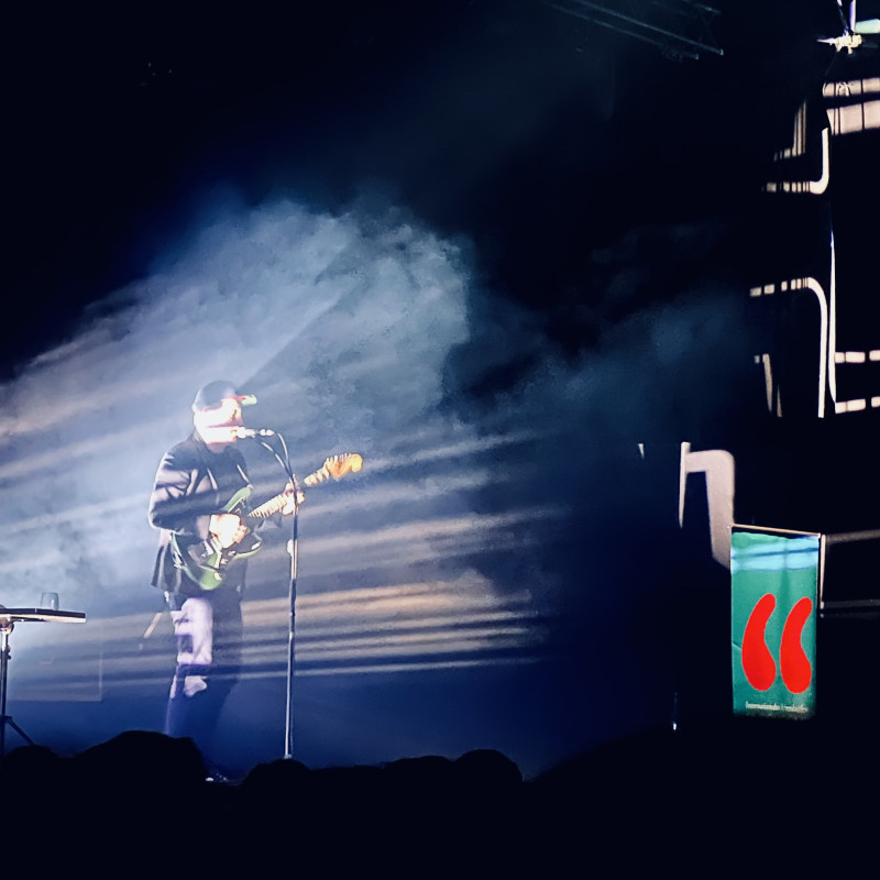 Thees Uhlmann steht mit Mütze und Gitarre auf der Bühne und wird von hinten angestrahlt. An der Wand entstehen durch das Licht abstrakte Muster. Neben ihm ein Banner mit dem Logo der Veranstaltung.