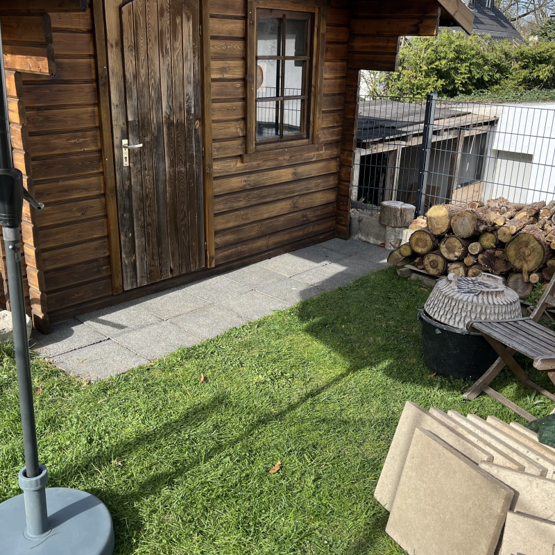 Graue Gehwegplatten liegen vor einem Gartenhaus, rechts ist ein Holzstapel zu sehen, daran sind die alten beigen Platten gelehnt.