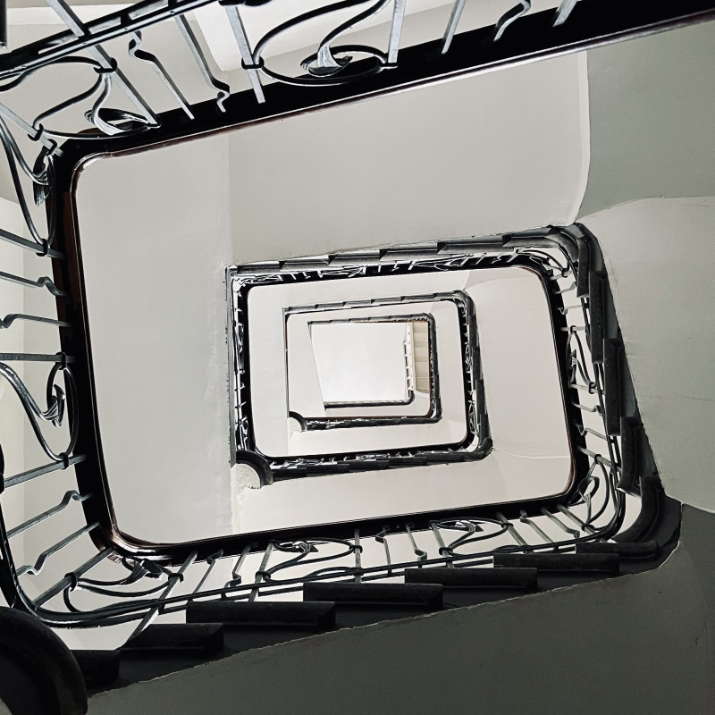 Blick von unten in ein gewundenes mehrstöckiges Treppenhaus. Das Geländer ist kunstvoll aus Metall gefertigt.