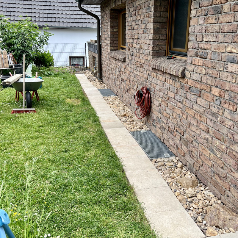 Auf einer grünen Wiese steht eine Schubkarre, an der ein Besen lehnt. Zwischen Wiese und dem Haus rechts daneben ist eine halbwegs gerade Fläche aus Gehwegplatten und grobem Kies.
