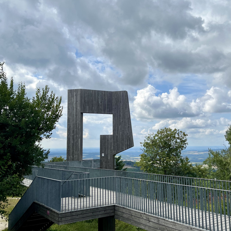Eine große Holzkonstruktion, zu der eine Brücke führt, steht auf einem Hügel vor dem Horizont.
