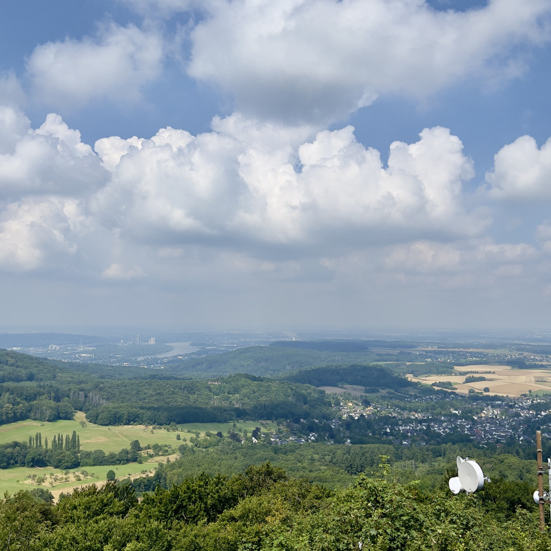 Blick vom Ölberg in Richtung Bonn. Ein Mittelgebirge mit Feldern und Orten, große Wolken am Himmel, im Hintergrund der Rhein.