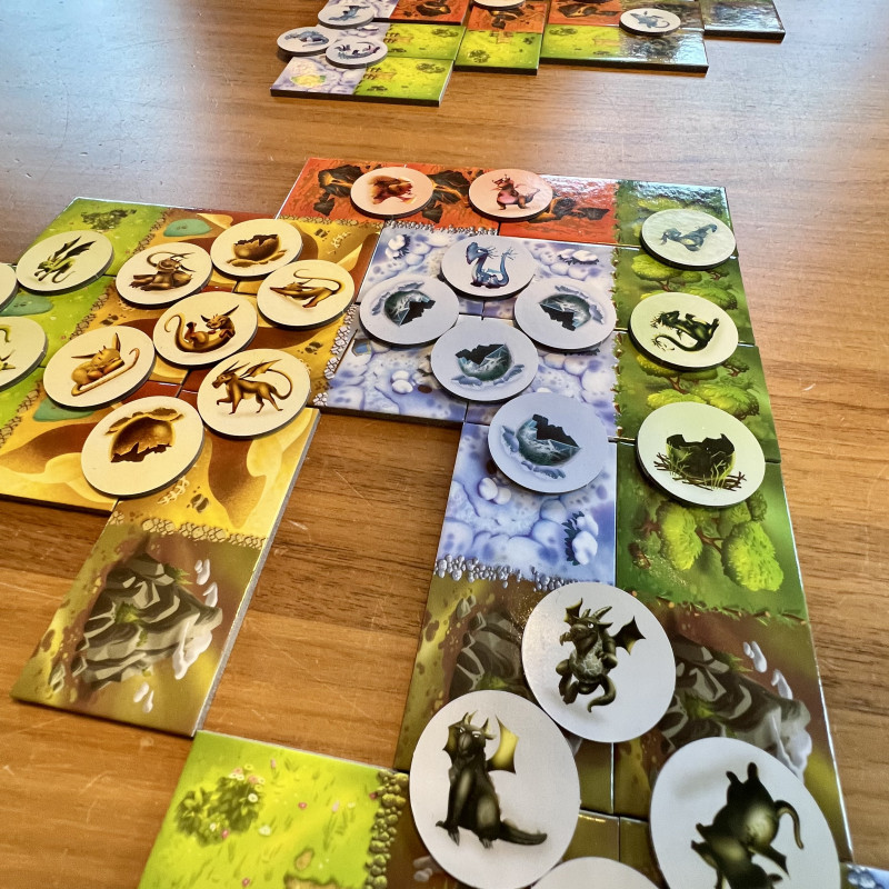 Auf einem Tisch liegen Spielkarten die eine Landschaft bilden. Darauf liegen Marken mit Drachensymbolen.